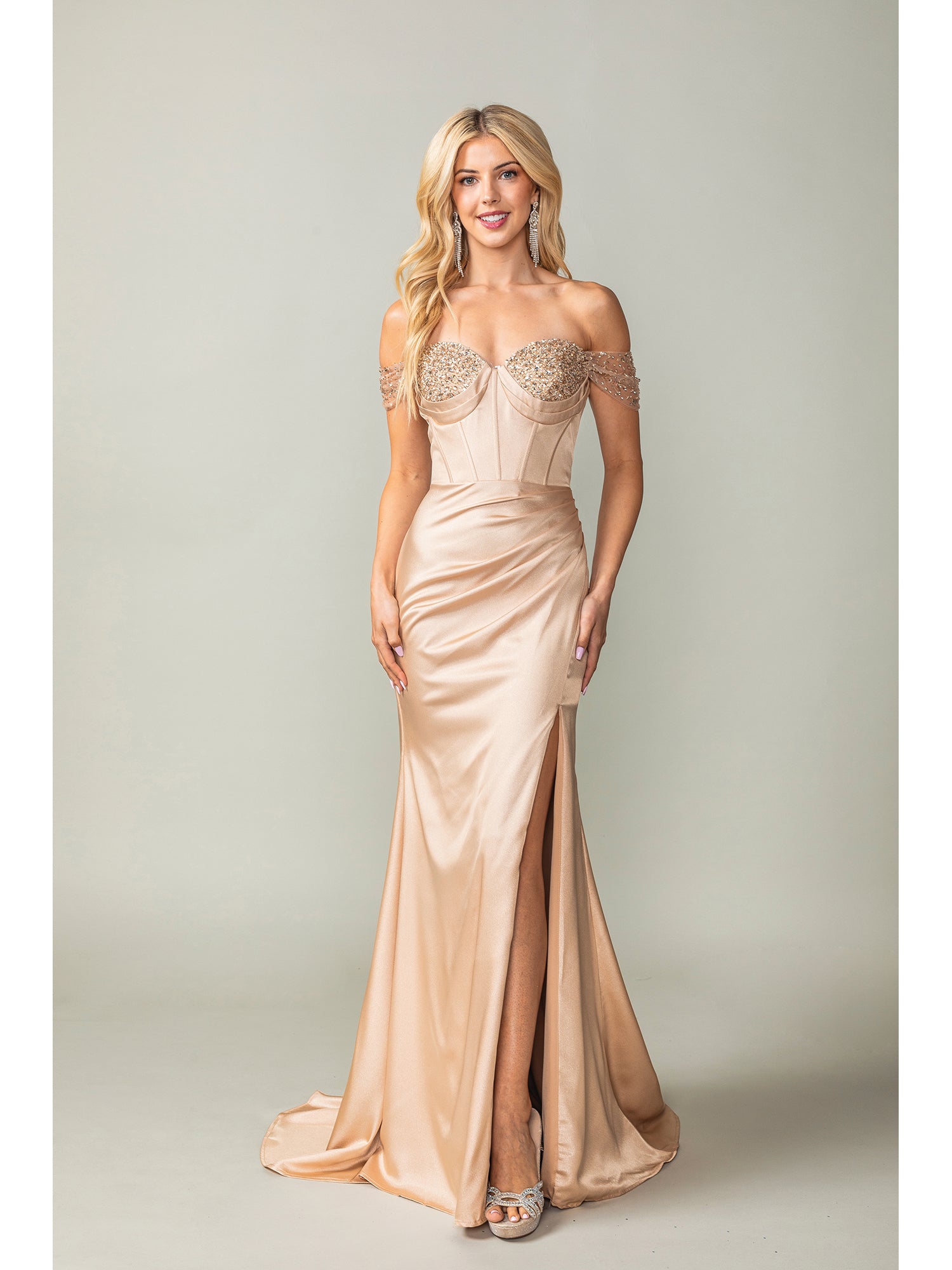 A woman in the Off-the-Shoulder Long Prom Dress 4401 with beaded details, a corset bodice, and thigh-high slit pairs the champagne gown with silver heels against a plain background.